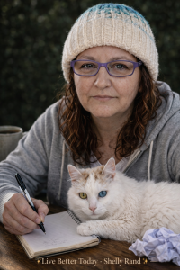 Shelly Rand with hazel eyes wearing a knit hat and glasses writes in a notebook at a wooden table while her white cat with one blue eye and one gold eye rests beside her; a coffee mug and crumpled paper sit nearby, with “Live Better Today – Shelly Rand” branding at the bottom.