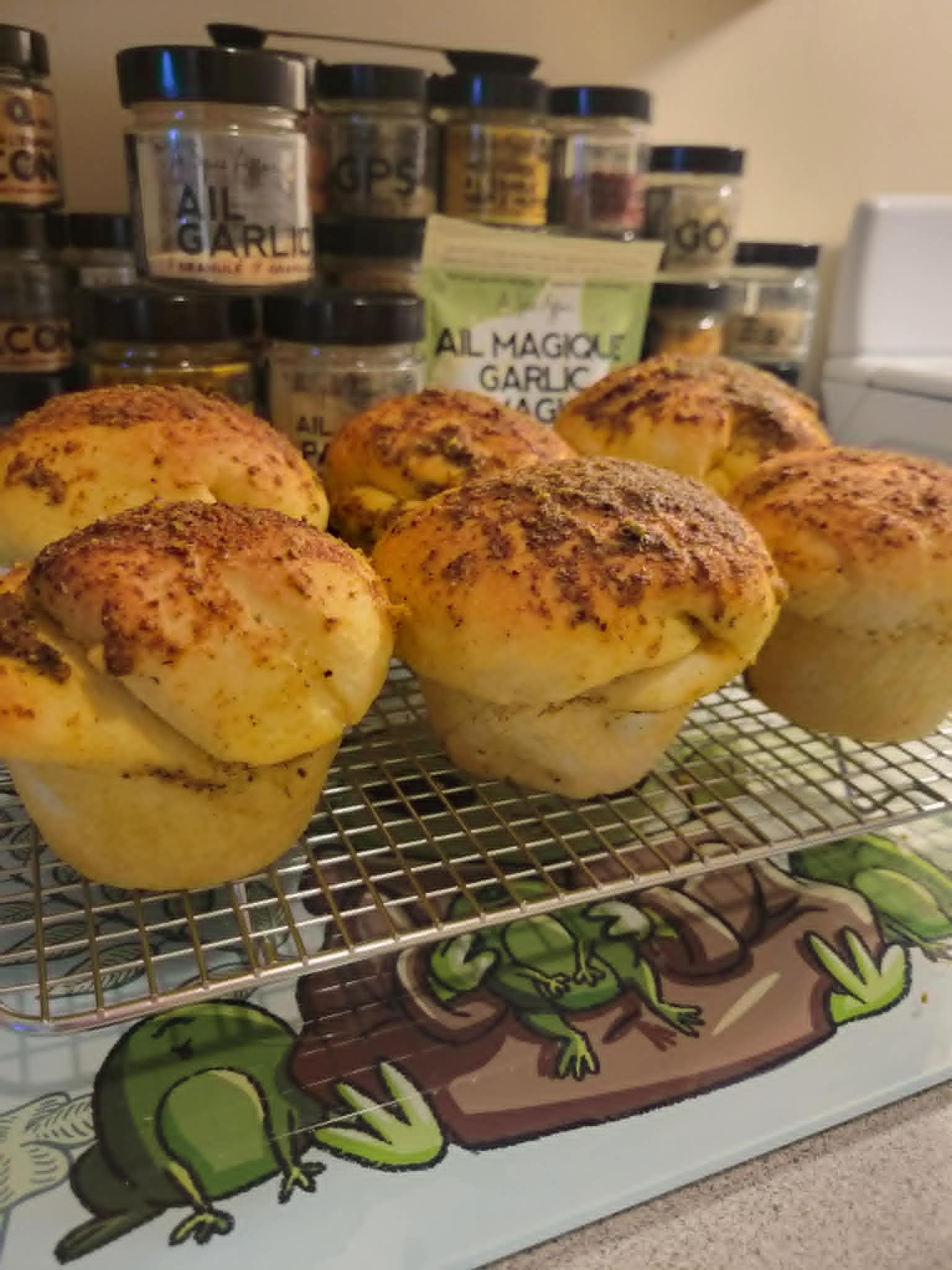 Homemade jumbo garlic parmesan buns cooling on a wire rack, with A Spice Affair Garlic Magic and other spices displayed behind them in Shellyβs kitchen.